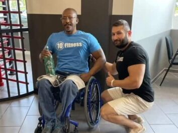 The image shows two men in a fitness setting. One man is seated in a blue wheelchair, wearing a light blue t-shirt with 