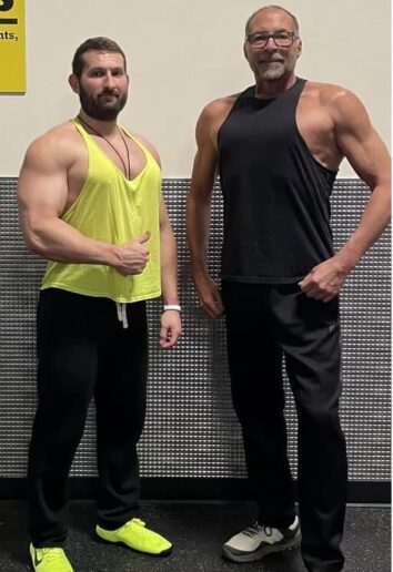 The image shows two men standing side by side in what appears to be a gym or fitness center. On the left is a younger man with a beard wearing a bright yellow tank top and black pants with neon yellow shoes. He's giving a thumbs up gesture. On the right is an older man with gray hair and glasses, wearing a black tank top and black pants. Both men are muscular and appear to be in good physical shape. They're posing in front of a metallic wall or partition. The overall impression is that of fitness enthusiasts or perhaps trainers at a gym.